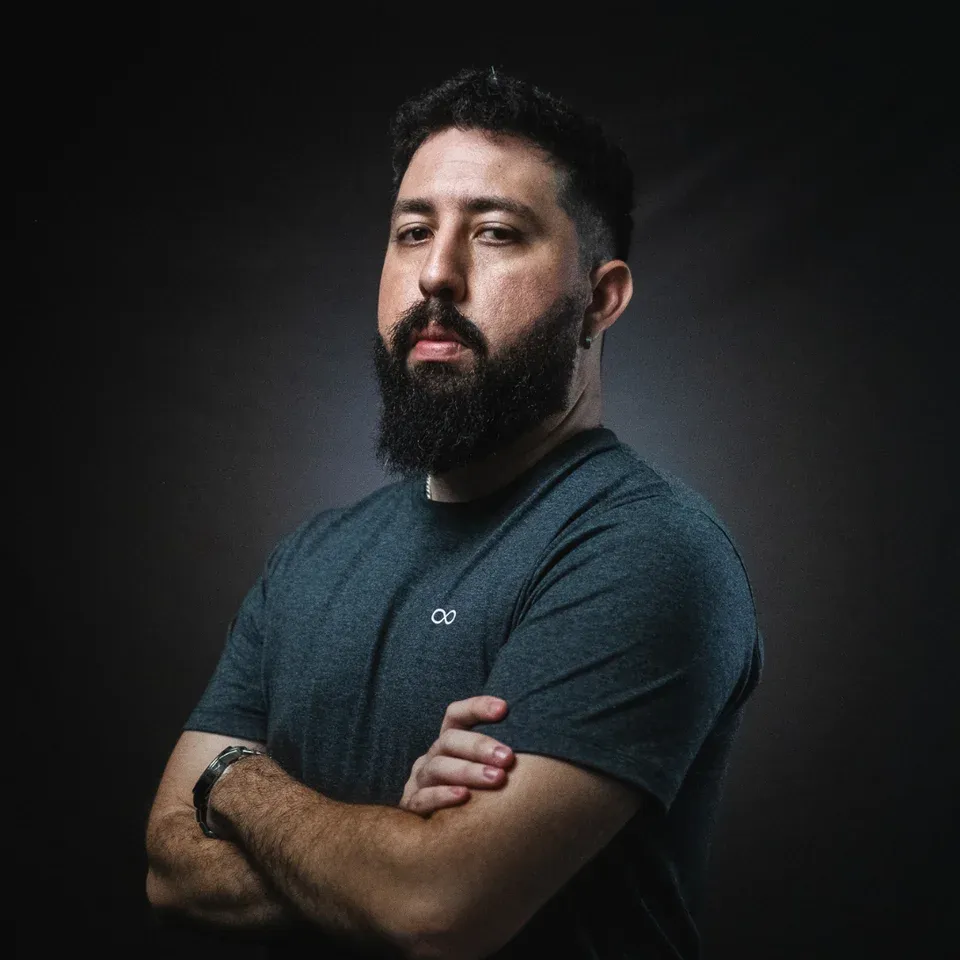 Marlon Maccedo, de camiseta escura e braços cruzados, em retrato com fundo escuro
